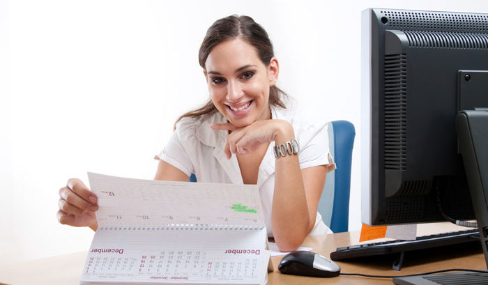young woman with pulled back brown hair smiles holding up a calendar at her desk