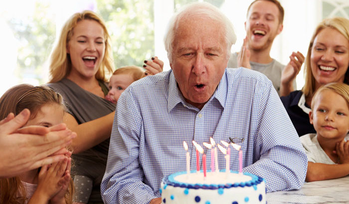 man turning sixty five blowing out birthday candles surrounded by friends and family