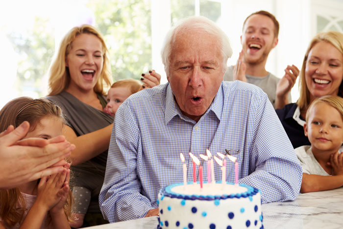 man turning sixty five blowing out birthday candles surrounded by friends and family