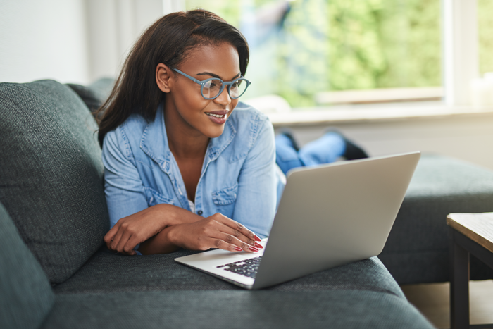 young african american woman on the couch wearing blue blocking eye glasses looking at her laptop